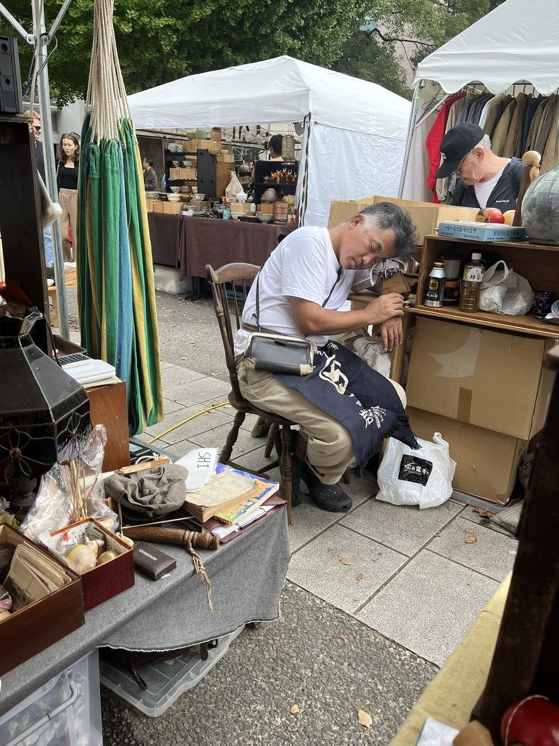 Een marktverkoper slaapt op een stoel tussen kraampjes met antiek en kleding op een vlooienmarkt