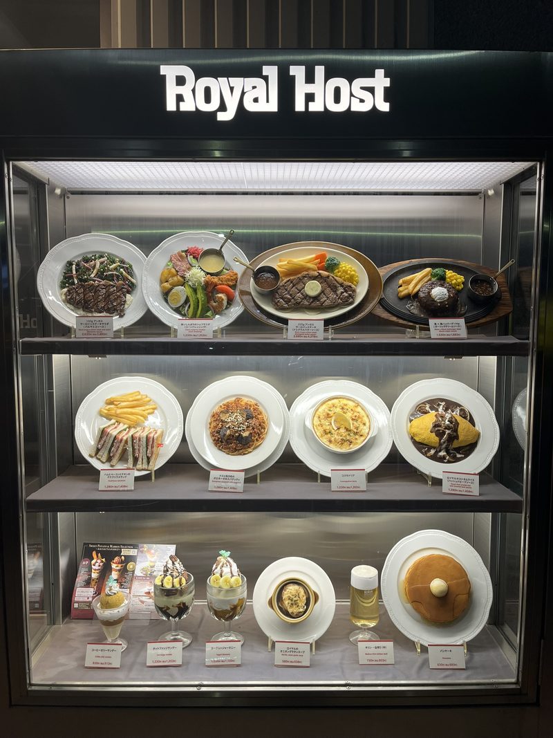 Een verlichte etalagekast van het restaurant Royal Host met plastic voorbeeldgerechten: steaks, pasta, desserts en pannenkoeken