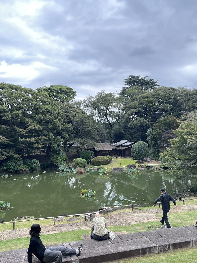 Een vijver met lotusbladeren in een Japanse tuin, met een traditioneel houten huis tussen de bomen en bezoekers op een stenen rand
